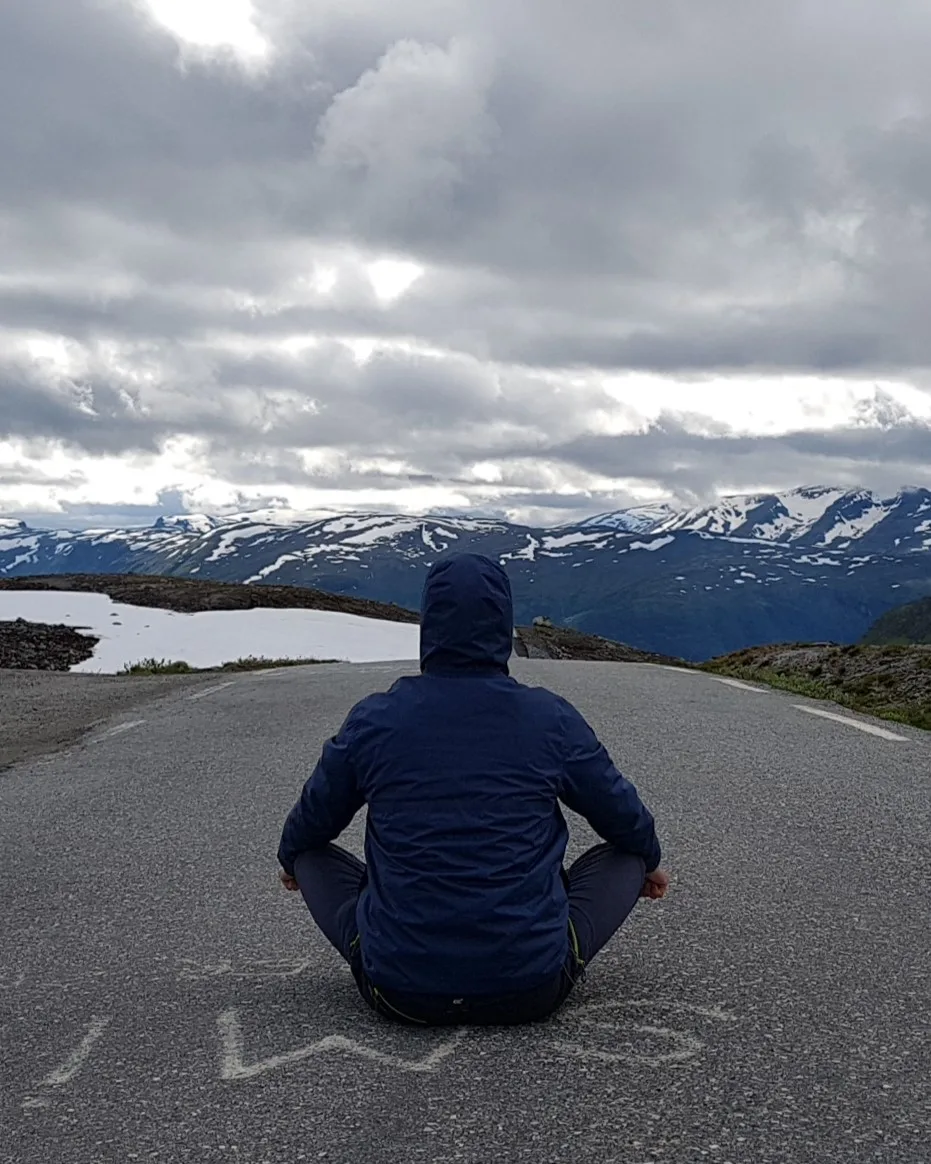 Jorge, de espaldas, sentado en mitad de una carretera de montaña con picos nevados al fondo.